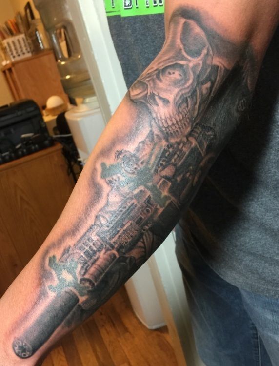 Tattoo of a skull and rifle on a person's forearm. Grayscale, detailed shading.
