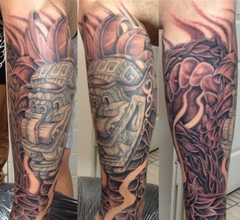 Arm tattoo of a detailed Mesoamerican mask surrounded by stylized leaves and patterns in black and gray.