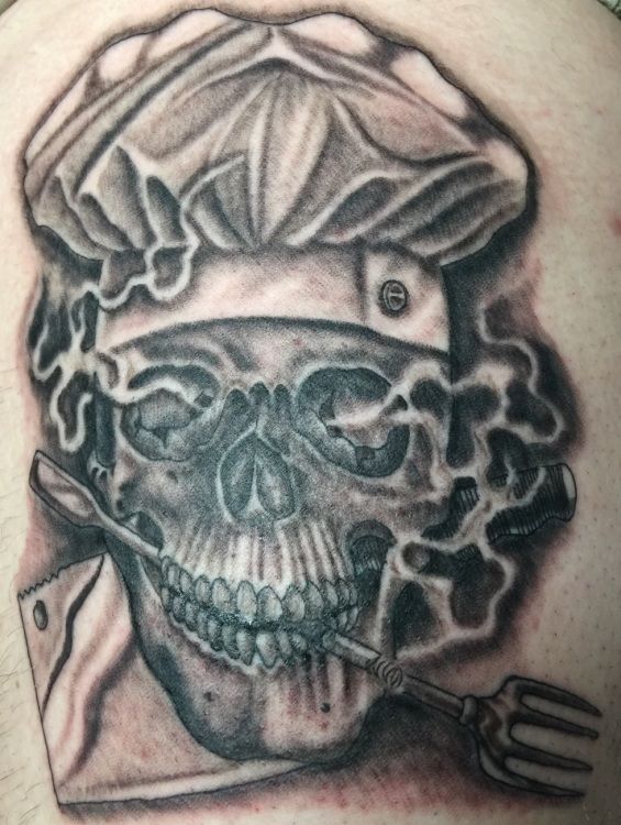 Skull wearing a chef's hat, holding a fork and knife, with a cloud of smoke. Black and grey tattoo.