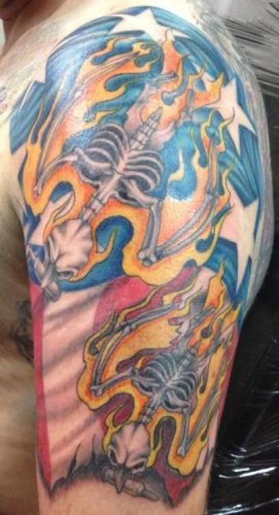 Tattoo on an arm: burning skeletal figures with a star field and flag-like design. Orange, blue, and red colors.