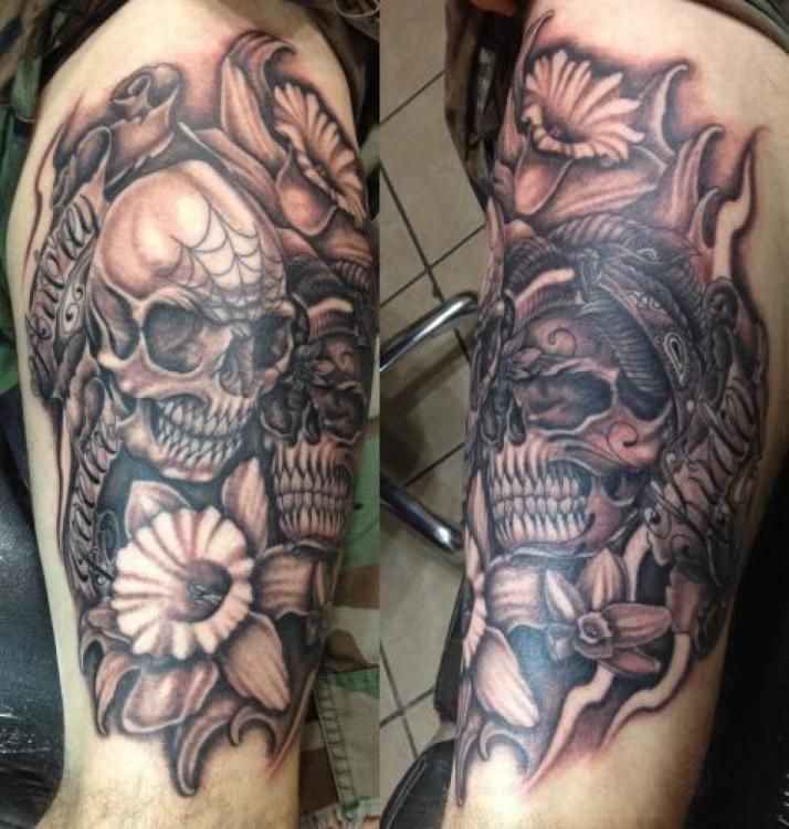 Tattoo on arm features two skulls, flowers, and foliage in grayscale.