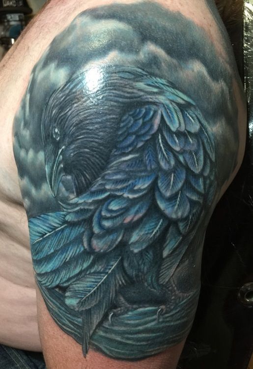 Raven tattoo on shoulder, blue and gray feathers, perched against stormy sky.