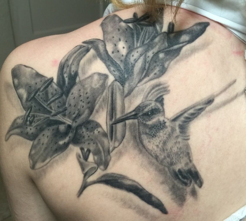 Black and gray tattoo of lilies and a hummingbird on a person's upper back.