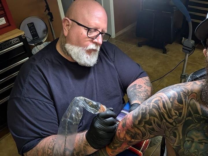 Tattoo artist with a gray beard and glasses giving a tattoo to someone's arm in a shop.
