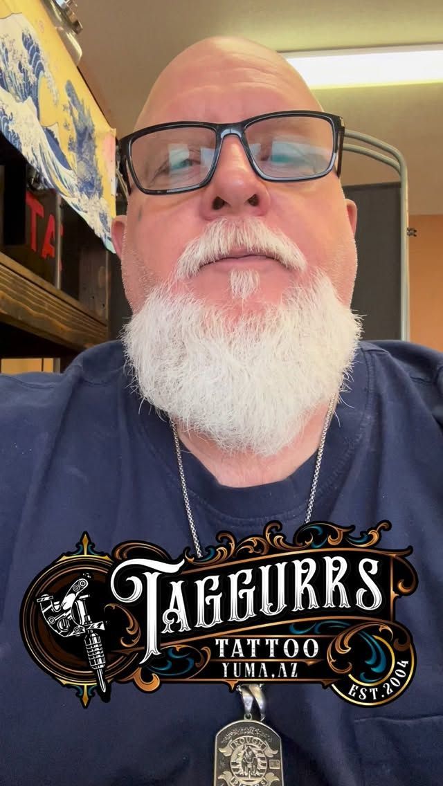 Man with glasses and white beard, wearing a blue shirt. The Taggurr's Tattoo Parlor logo is overlaid.