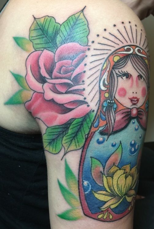 Tattoo on arm of a Russian nesting doll and a pink rose, with green leaves, and a yellow flower.
