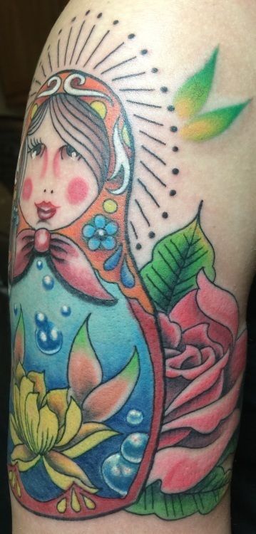 Colorful tattoo on an arm, featuring a Matryoshka doll, a rose, and floral elements.