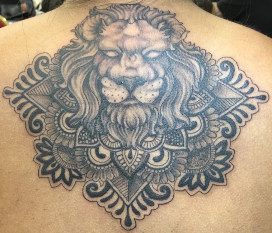 Tattoo of a lion's face with a detailed mandala design on a person's back; grayscale.