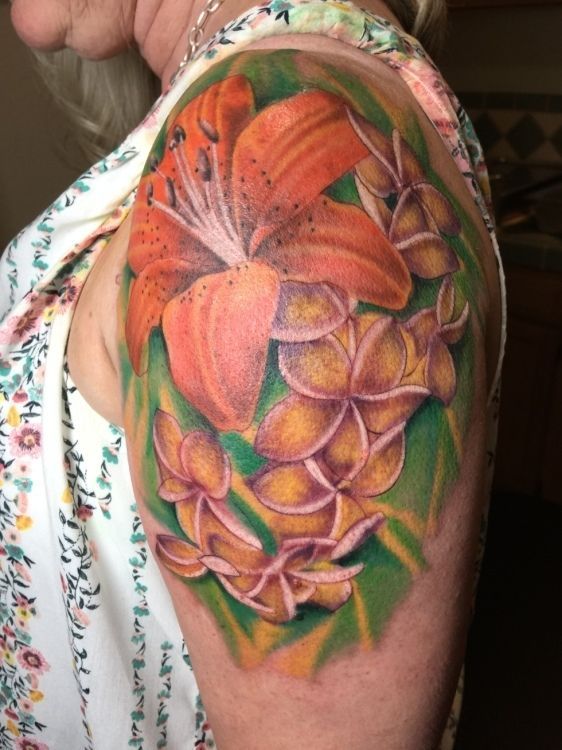 Tattoo of orange lily and yellow plumeria flowers on a person's upper arm, with green leaves and a floral shirt.