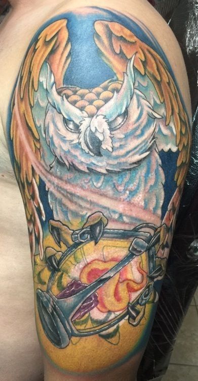 Colorful owl tattoo on a shoulder and upper arm, with wings, an hourglass, and blue/yellow hues.