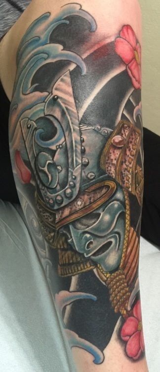 Arm tattoo of a samurai helmet with a mask, waves, and flowers; blue, gray, gold, and red hues.