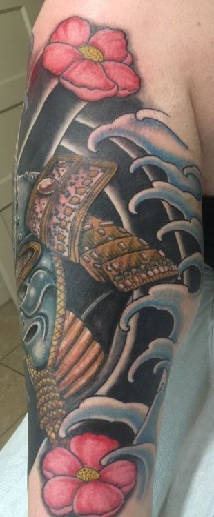 Tattoo of a samurai helmet with flowers and water waves in dark blue, red, and gold colors on an arm.