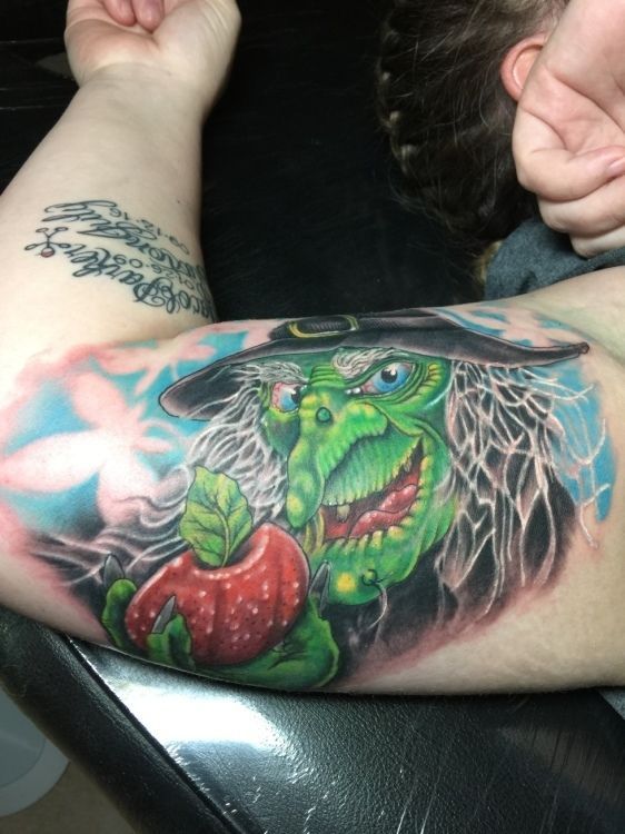 Tattoo of a green witch with a black hat holding a red apple; blue and white background.