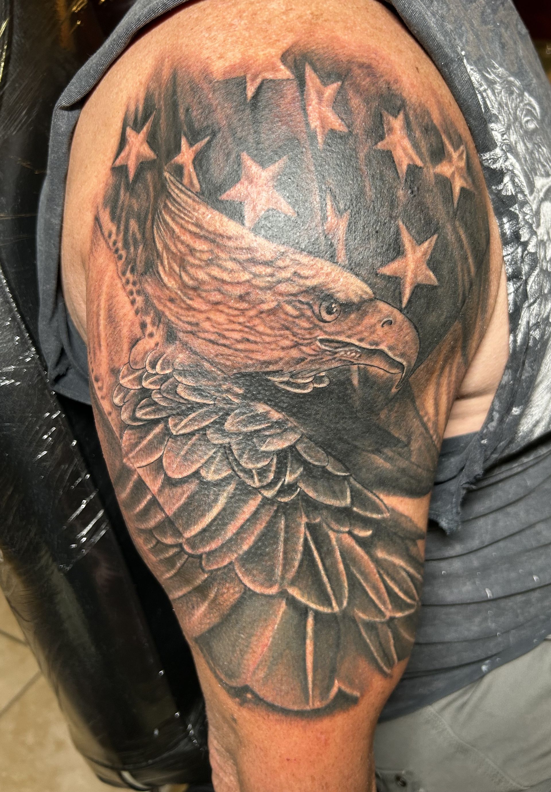 Tattoo of an eagle with its head and wing detailed on a shoulder. Stars and a dark background are also present.