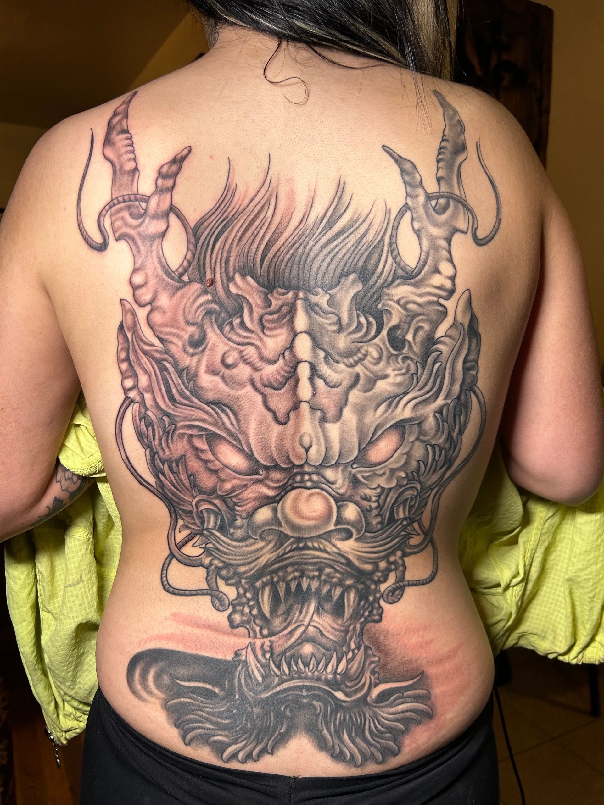 Back tattoo of a detailed, gray and red-toned demonic face with horns and open mouth.