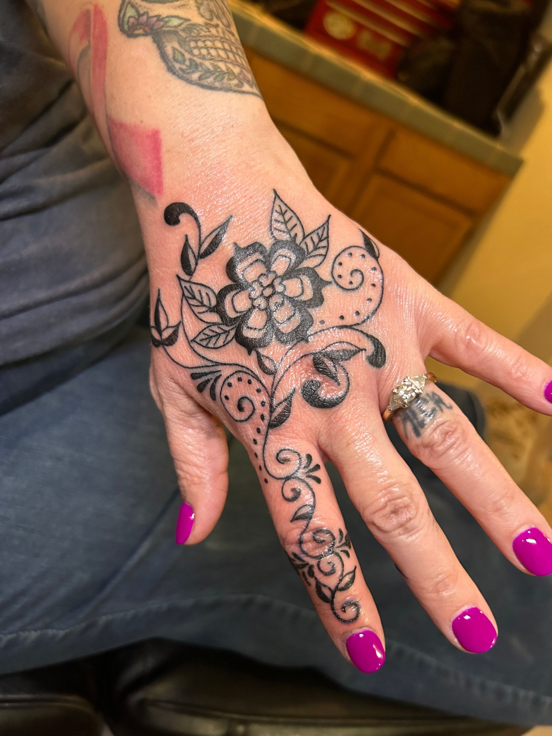 Hand tattoo of a flower and vines. The hand has bright pink nail polish and a ring.
