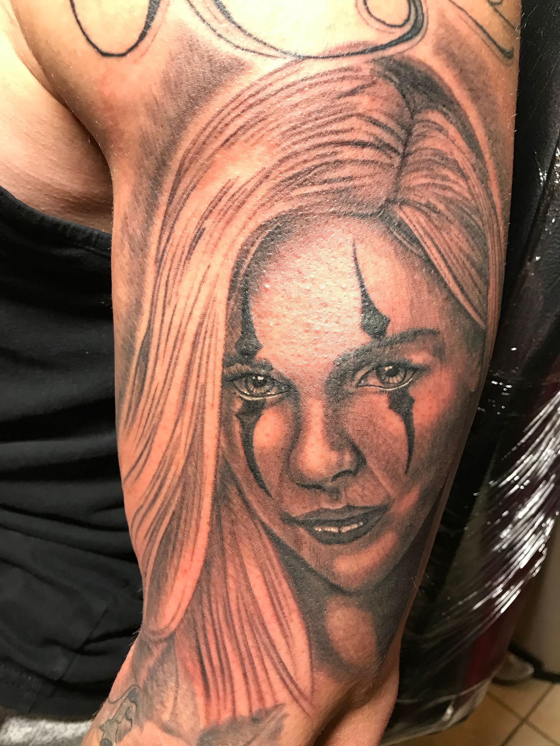 Tattoo of a person's face with long blonde hair and clown-like eye makeup on an arm.