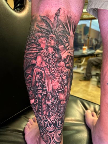 Detailed black and gray tattoo on a leg depicting a figure with headdress, surrounding structures, and sunburst.