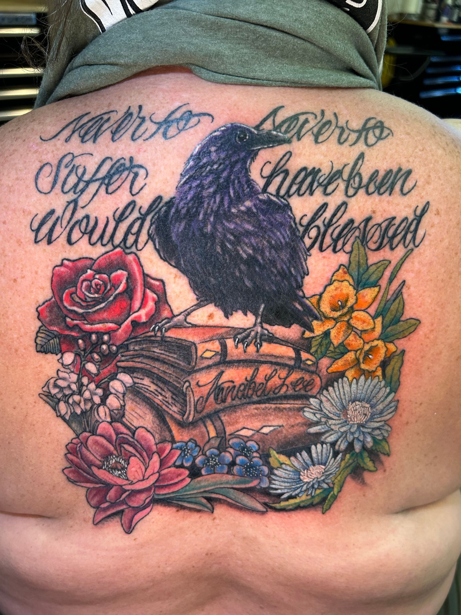 Tattoo of a black raven on books surrounded by flowers; script reads, 