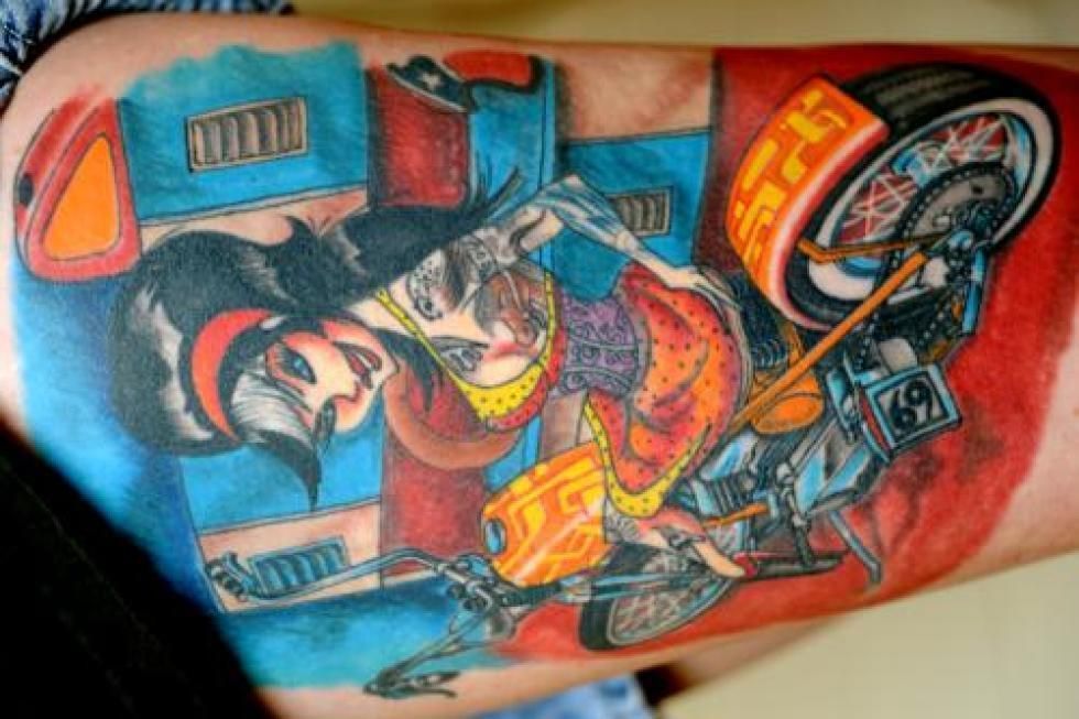 Tattoo of a woman riding a motorcycle with an orange wheel, on a red and blue background.