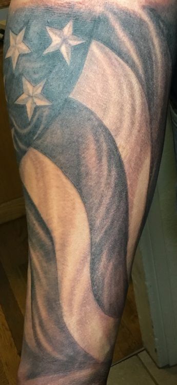 A black and white tattoo of a waving American flag on an arm. Three stars are visible in the top left corner.