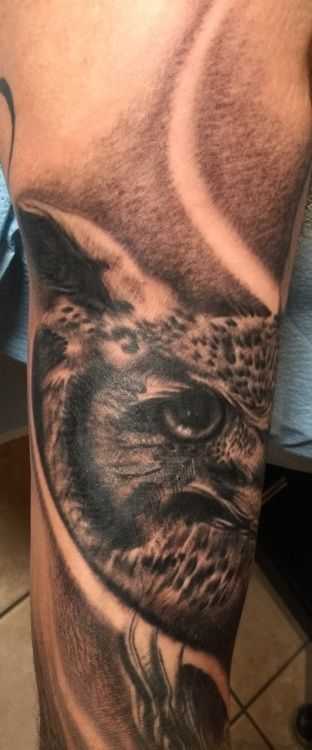 Close-up of an owl eye tattoo on a person's arm, shaded in black and gray.