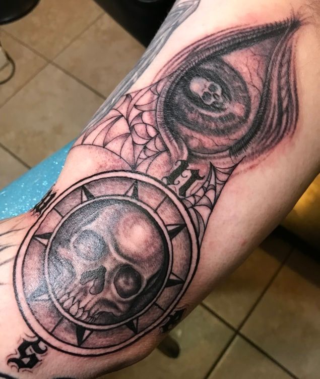 Tattoo on arm features a skull in a compass-like design and a swirling eye with a skull inside; cobweb detail.