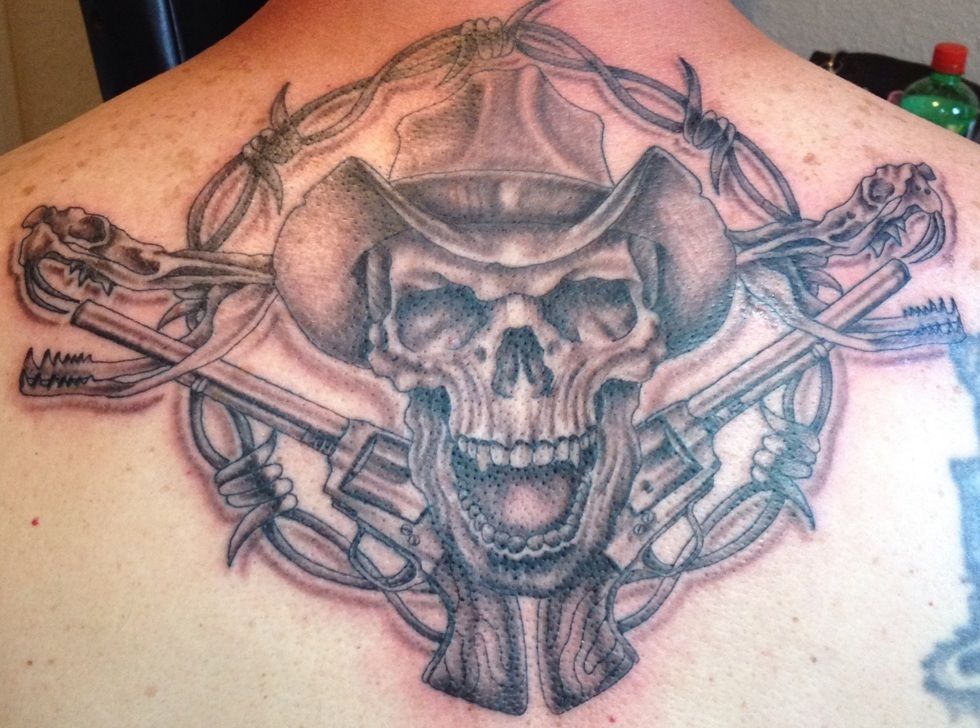 Tattoo of a skull wearing a cowboy hat, crossed revolvers, and barbed wire on a back.
