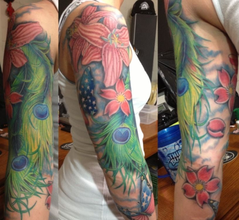 Tattoo of a woman's arm with colorful flowers, peacock feathers, and a partial American flag.
