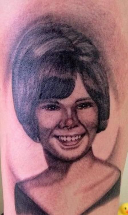 Portrait tattoo of a smiling woman with bouffant hair. Black and gray shading, with a dark background.