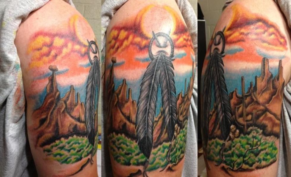 Tattoo of a landscape with feathers and mountains under a colorful sunset.
