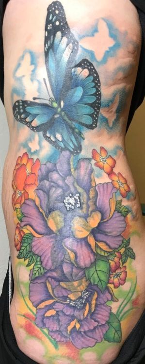 A tattoo on a person's side with a blue butterfly, purple and orange flowers, and a blue sky background.