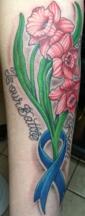 Tattoo of pink daffodils, green stems, and a blue ribbon with text on a person's arm.