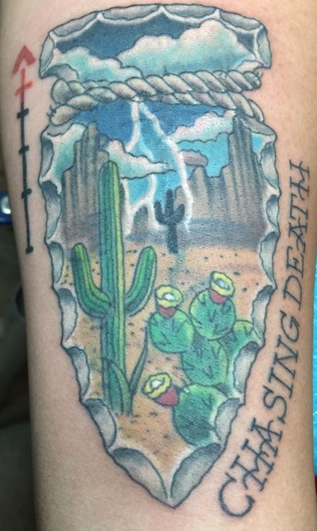 Tattoo of desert landscape inside a gray arrowhead shape. 