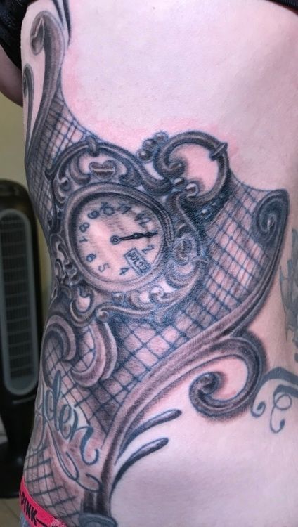 Tattoo of a pocket watch within ornate framing on a person's side, with a lace-like background.