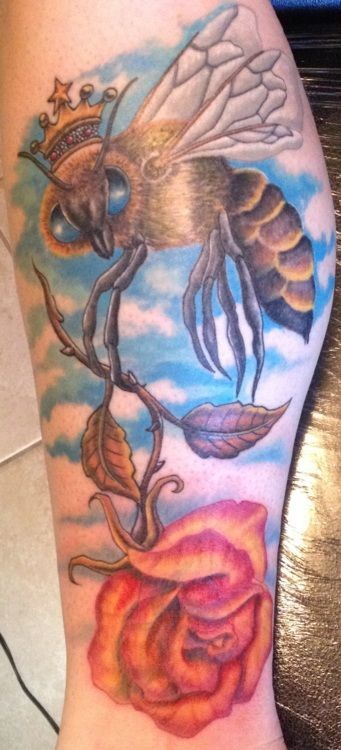 Tattoo of a queen bee wearing a crown, perched on a rose vine, against a blue sky background.