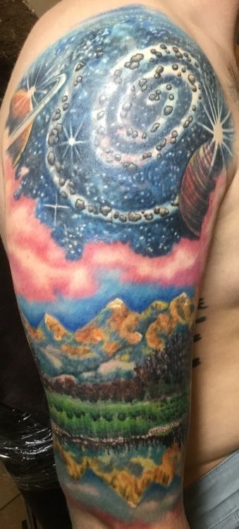 Tattoo on arm depicts a vibrant cosmic scene with swirling galaxies, planets, and a mountainous landscape.