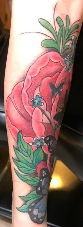 Tattoo of a large red flower with green leaves, blue flowers, and black berries on a forearm.