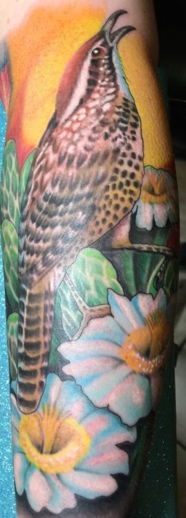 Tattoo of a bird on a branch, with flowers in the foreground. Yellow and blue colors.