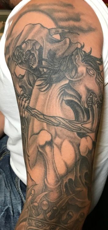 Tattoo of a figure with skeletal features holding a branch, surrounded by stylized details on an arm.