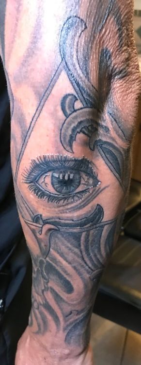 Tattoo of a detailed eye within a triangle, surrounded by ornate swirls, on a person's arm.