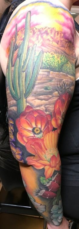 Full arm tattoo featuring a desert landscape with a cactus, vibrant orange flowers, and a sunset.