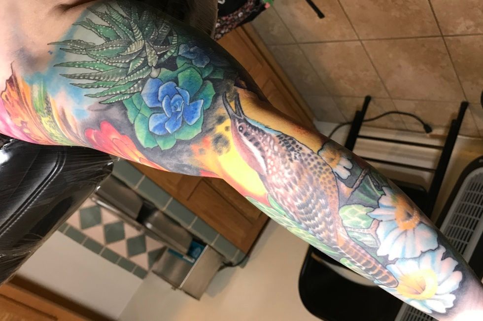 Colorful arm tattoo: bird, flowers, foliage, blue, green, yellow, on a human arm.