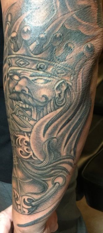 Tattoo of a stylized face wearing a crown, with swirling designs and shading.