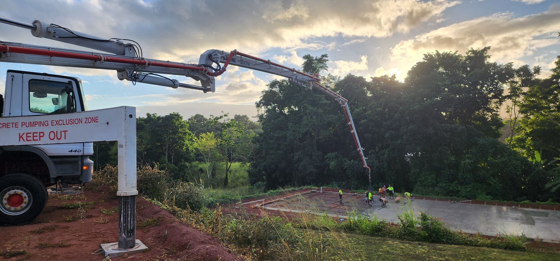 Water truck spraying a stream onto a grassy roadside at sunset, with trees and hills in the background