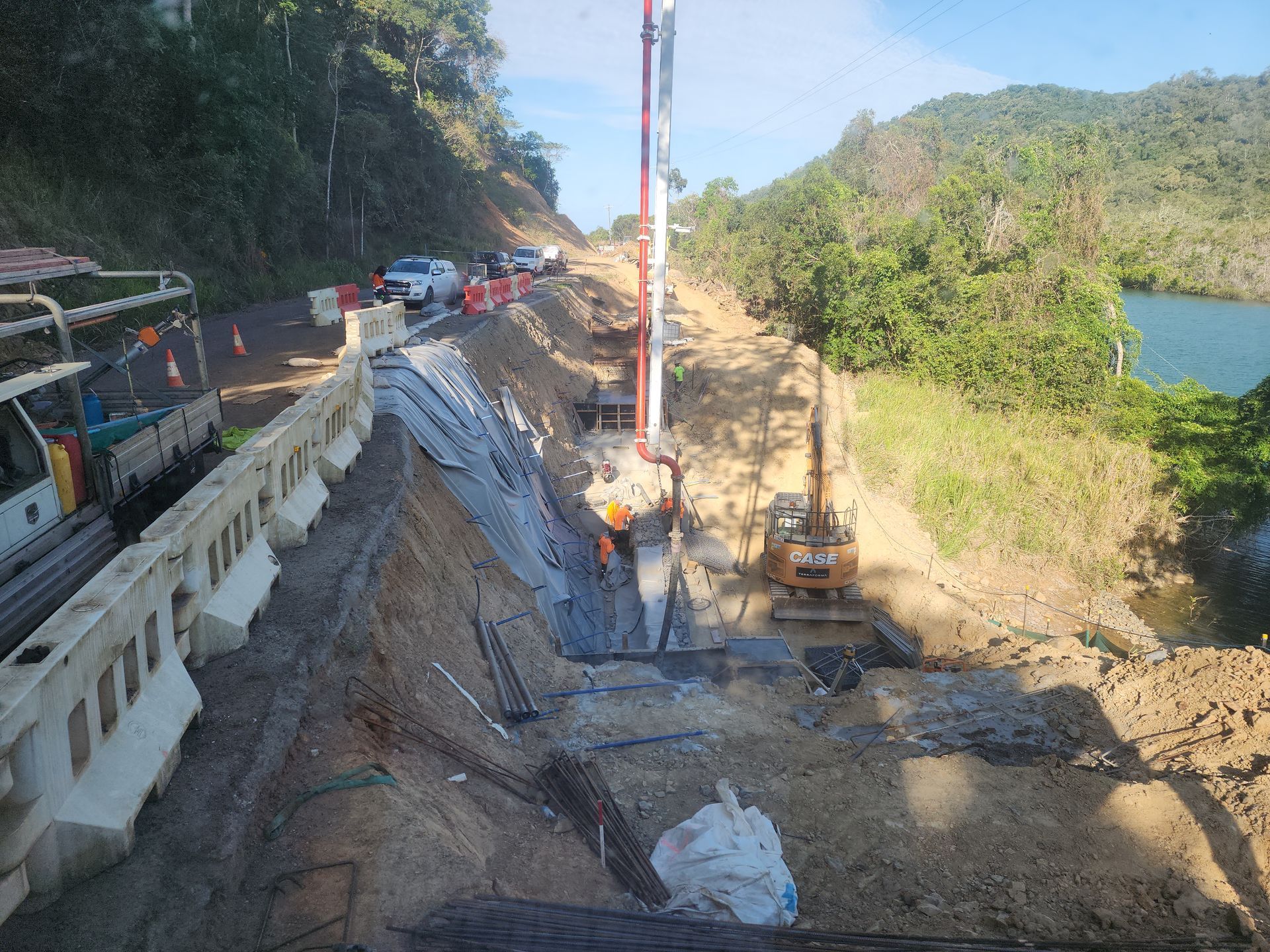 Roadwork on a hillside beside a river, with excavators, concrete barriers, and a steep dirt slope.