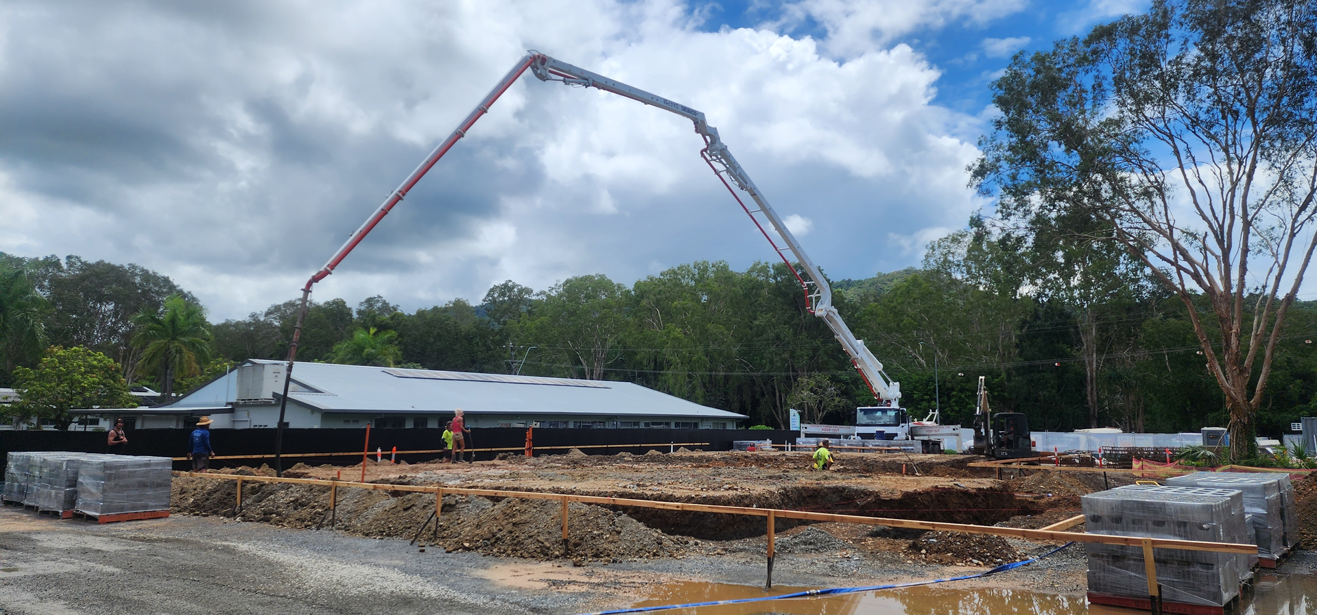 Construction site with a concrete pump boom arcing over a low building under a cloudy sky
