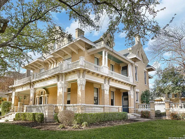 Historic beige Victorian house with ornate white trim and wraparound porch under blue sky.
