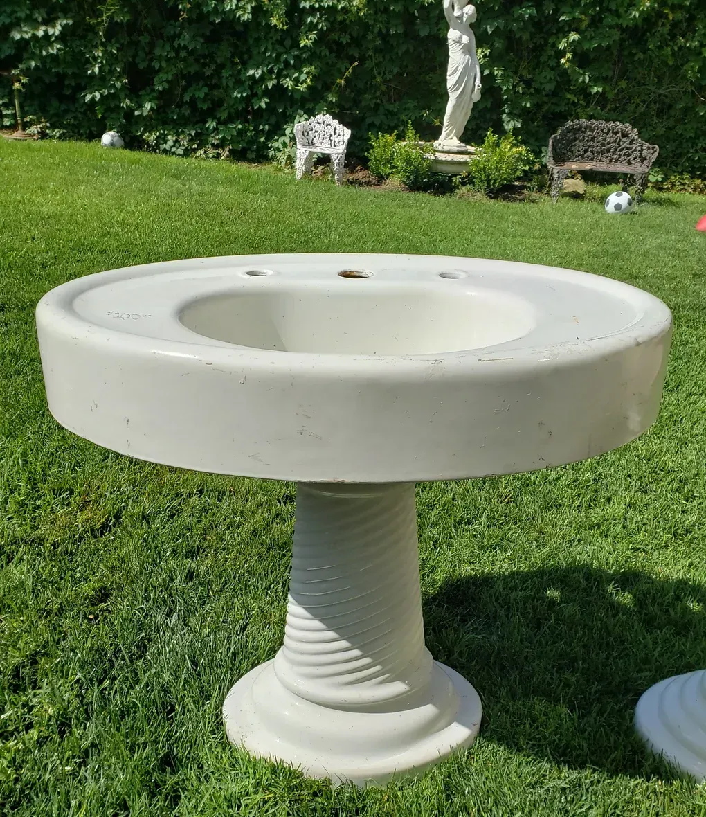 A white pedestal sink sits in the middle of a lush green field
