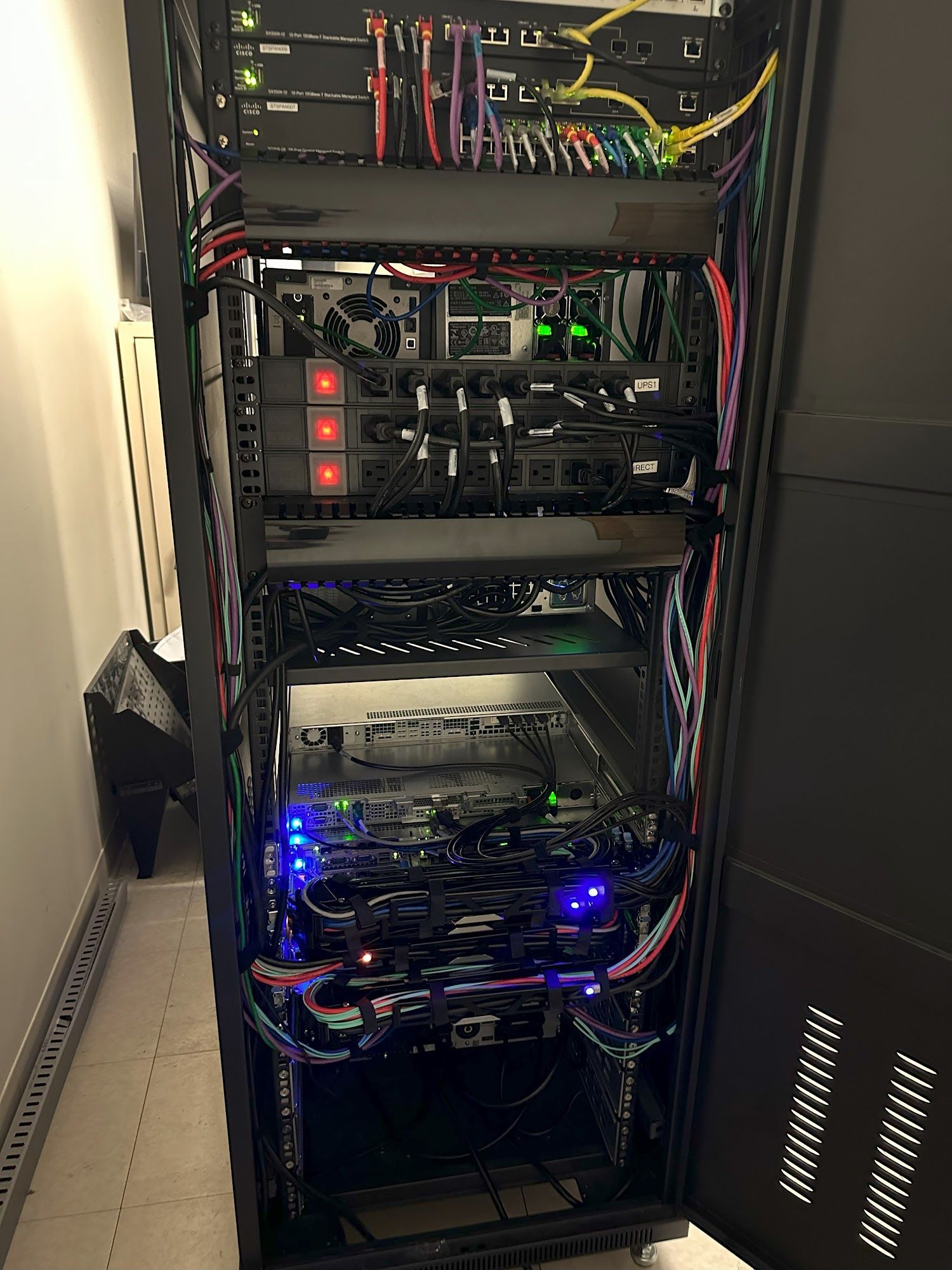 Server rack with various electronic devices, cables, and blinking lights. Dark setting, organized appearance.